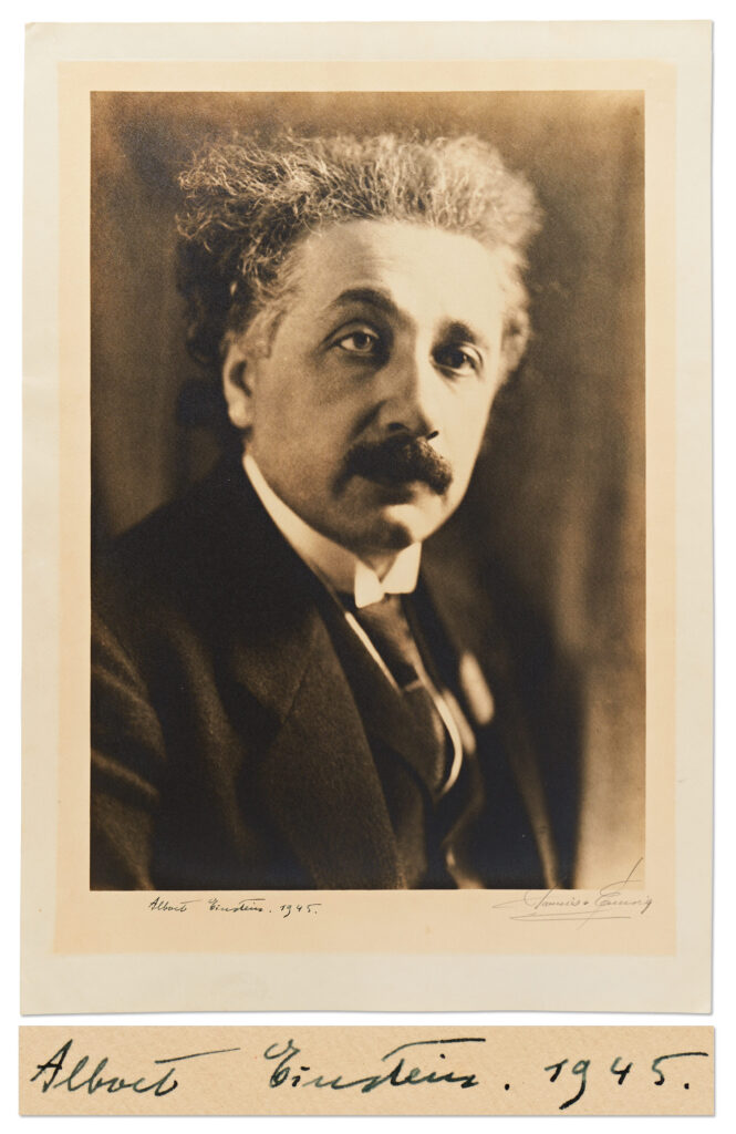 Black and white portrait of Albert Einstein wearing a suit jacket and tie. Photo has thick borders and Einstein signs along bottom margin.