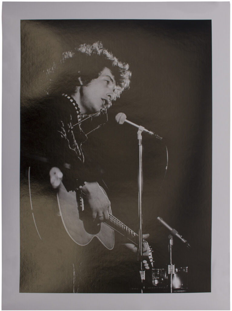 Bob Dylan performing with a guitar in front of a mic while wearing a harmonica holder.