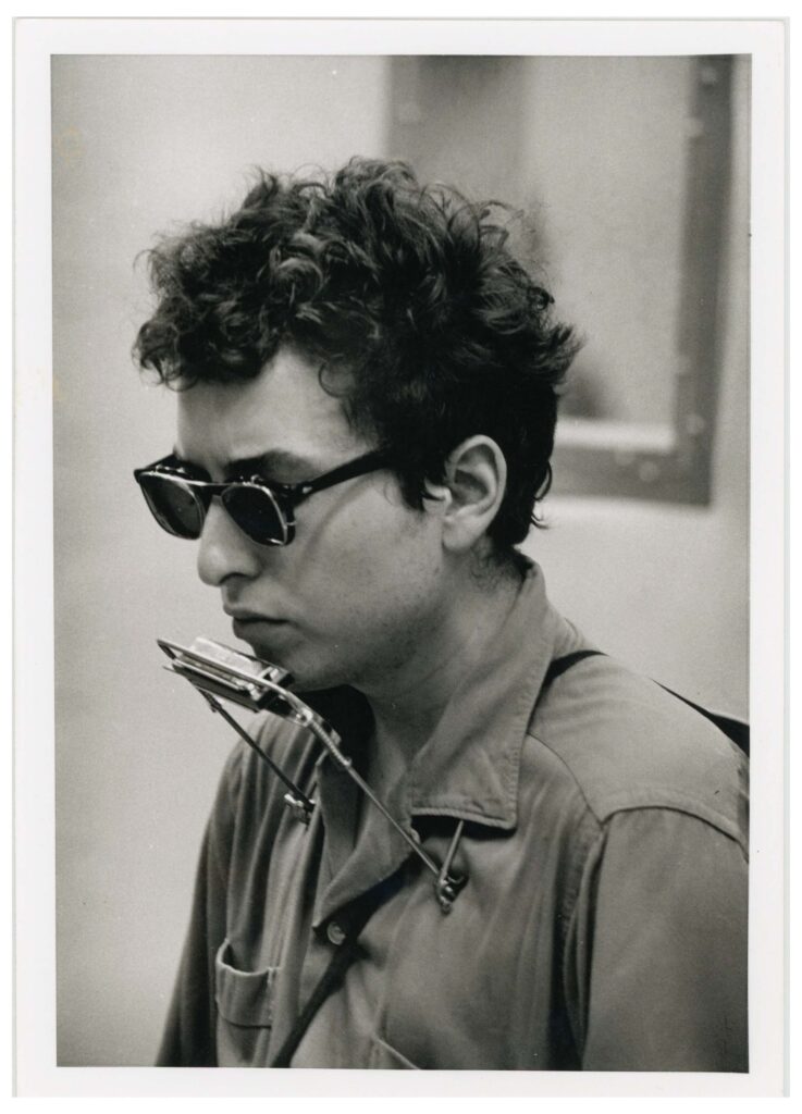 Bob Dylan wearing a collared shirt, sunglasses and a harmonica holder on neck.